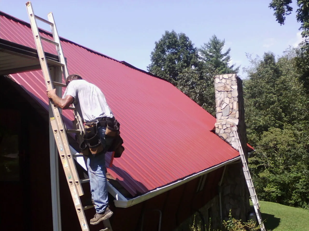 Roofer on a ladder beside a red metal roof and stone chimney — Soffit & Fascia Repair by Roofing Brothers in Waynoka